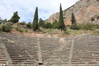 An ancient stone amphitheater set into a hillside is surrounded by tall cypress trees and rocky terrain, blending natural and historical elements.