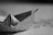 A black and white image featuring a paper boat made from a printed newspaper or similar text material. The background appears to be a blurred map or possibly a desk surface with text partially visible.