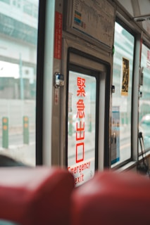 The image captures the interior of a vehicle, likely a bus or train, focusing on the emergency exit door. Prominently displayed on the door is a sign in both red Japanese characters and English that reads 'Emergency Exit'. The background features windows through which an urban environment is partially visible, with structures and blurred vehicles outside. Red seats are partially visible in the foreground, adding to the sense of being within a passenger vehicle.