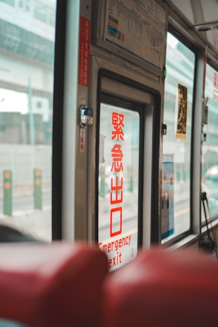 The image captures the interior of a vehicle, likely a bus or train, focusing on the emergency exit door. Prominently displayed on the door is a sign in both red Japanese characters and English that reads 'Emergency Exit'. The background features windows through which an urban environment is partially visible, with structures and blurred vehicles outside. Red seats are partially visible in the foreground, adding to the sense of being within a passenger vehicle.