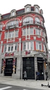 A large, ornate building with a bright red facade and intricate architectural details, including numerous windows with decorative frames and iron balconies. The ground level features retail store signs for 'Rolex' and 'Marcolino,' and the walls are adorned with colorful floral decorations. Two people are walking on the sidewalk below, and a scooter is visible nearby.
