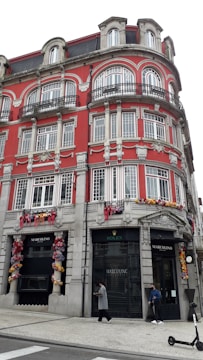 A large, ornate building with a bright red facade and intricate architectural details, including numerous windows with decorative frames and iron balconies. The ground level features retail store signs for 'Rolex' and 'Marcolino,' and the walls are adorned with colorful floral decorations. Two people are walking on the sidewalk below, and a scooter is visible nearby.