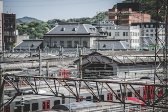 Urban scene featuring a railway station surrounded by various buildings. Trains are visible on the tracks, and overhead electrical infrastructure is present. The architecture includes a mix of modern and older-style buildings, with greenery visible in the background.