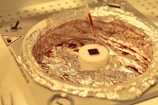 A laboratory setup featuring a small square object placed on a circular white platform wrapped with aluminum foil. Above it, a pipette drips a red liquid onto the platform. The background consists of a laboratory environment with indicators and instructions visible.