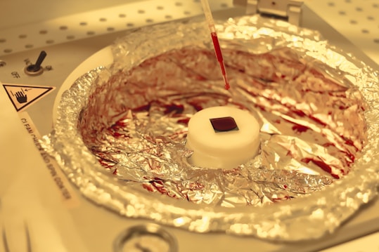 A laboratory setup featuring a small square object placed on a circular white platform wrapped with aluminum foil. Above it, a pipette drips a red liquid onto the platform. The background consists of a laboratory environment with indicators and instructions visible.