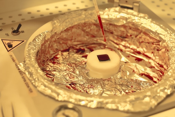 A laboratory setup featuring a small square object placed on a circular white platform wrapped with aluminum foil. Above it, a pipette drips a red liquid onto the platform. The background consists of a laboratory environment with indicators and instructions visible.