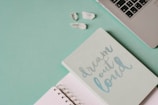 A soft green notebook with the phrase 'dream out loud' in light blue lettering lies next to an open lined notebook. In the top right corner, part of a silver laptop keyboard is visible, and a few clear quartz crystals rest nearby on a green surface.