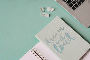 A soft green notebook with the phrase 'dream out loud' in light blue lettering lies next to an open lined notebook. In the top right corner, part of a silver laptop keyboard is visible, and a few clear quartz crystals rest nearby on a green surface.
