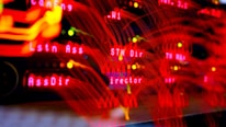 A close-up view of a server room or data center with bright red LED lights. The lights form various technical terms, suggesting network or server activity. The background is dark, highlighting the vibrant, dynamic lines and letters.