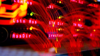 A close-up view of a server room or data center with bright red LED lights. The lights form various technical terms, suggesting network or server activity. The background is dark, highlighting the vibrant, dynamic lines and letters.
