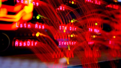 A close-up view of a server room or data center with bright red LED lights. The lights form various technical terms, suggesting network or server activity. The background is dark, highlighting the vibrant, dynamic lines and letters.