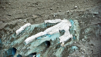 A glacier partially covered with white protective sheets is surrounded by rugged, rocky terrain. The ice beneath the sheets displays shades of blue and green, indicating its glacial nature. The area appears desolate and isolated.