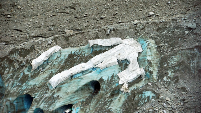 A glacier partially covered with white protective sheets is surrounded by rugged, rocky terrain. The ice beneath the sheets displays shades of blue and green, indicating its glacial nature. The area appears desolate and isolated.