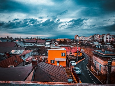 An urban neighborhood scene with densely packed houses and buildings under a dramatic, cloudy sky. A vibrant orange house stands out among the more muted tones of its surroundings. The winding road leads through the residential area, where a few parked cars can be seen. The distant skyline features taller apartment buildings.