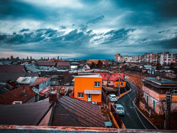 An urban neighborhood scene with densely packed houses and buildings under a dramatic, cloudy sky. A vibrant orange house stands out among the more muted tones of its surroundings. The winding road leads through the residential area, where a few parked cars can be seen. The distant skyline features taller apartment buildings.