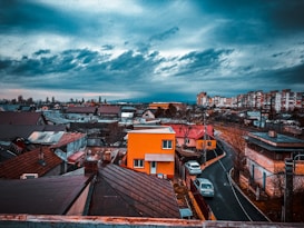 An urban neighborhood scene with densely packed houses and buildings under a dramatic, cloudy sky. A vibrant orange house stands out among the more muted tones of its surroundings. The winding road leads through the residential area, where a few parked cars can be seen. The distant skyline features taller apartment buildings.