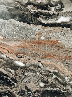 Intricate marble texture with swirling patterns of gray, black, and earthy brown tones. The surface features various layers and formations, highlighting the natural veining typical of stone.