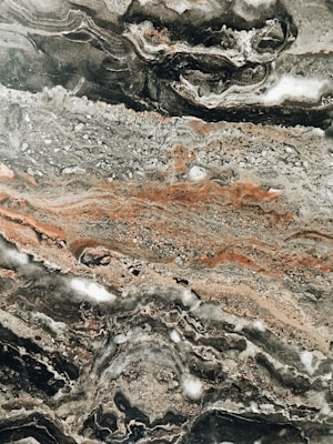 Intricate marble texture with swirling patterns of gray, black, and earthy brown tones. The surface features various layers and formations, highlighting the natural veining typical of stone.