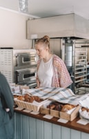 A woman stands behind a bakery counter, looking at an array of pastries and baked goods laid out in baskets and trays, covered partially by a checkered cloth. She appears to be in a bakery shop with large ovens and baking equipment visible in the background.
