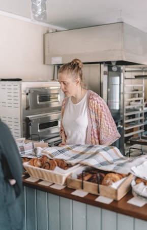 A woman stands behind a bakery counter, looking at an array of pastries and baked goods laid out in baskets and trays, covered partially by a checkered cloth. She appears to be in a bakery shop with large ovens and baking equipment visible in the background.
