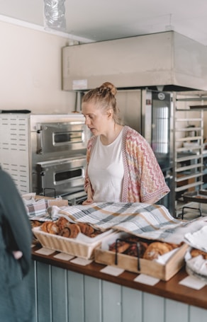 A woman stands behind a bakery counter, looking at an array of pastries and baked goods laid out in baskets and trays, covered partially by a checkered cloth. She appears to be in a bakery shop with large ovens and baking equipment visible in the background.