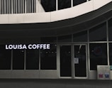 A storefront featuring large glass windows and a sign displaying 'Louisa Coffee' in bright, bold letters. The entrance consists of glass doors with signage posted on them. The exterior design is modern, with clean lines and a minimalist aesthetic. Two recycling bins are positioned to the right of the entrance.