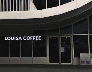 A storefront featuring large glass windows and a sign displaying 'Louisa Coffee' in bright, bold letters. The entrance consists of glass doors with signage posted on them. The exterior design is modern, with clean lines and a minimalist aesthetic. Two recycling bins are positioned to the right of the entrance.