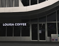 A storefront featuring large glass windows and a sign displaying 'Louisa Coffee' in bright, bold letters. The entrance consists of glass doors with signage posted on them. The exterior design is modern, with clean lines and a minimalist aesthetic. Two recycling bins are positioned to the right of the entrance.