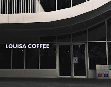 A storefront featuring large glass windows and a sign displaying 'Louisa Coffee' in bright, bold letters. The entrance consists of glass doors with signage posted on them. The exterior design is modern, with clean lines and a minimalist aesthetic. Two recycling bins are positioned to the right of the entrance.