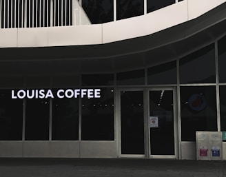 A storefront featuring large glass windows and a sign displaying 'Louisa Coffee' in bright, bold letters. The entrance consists of glass doors with signage posted on them. The exterior design is modern, with clean lines and a minimalist aesthetic. Two recycling bins are positioned to the right of the entrance.