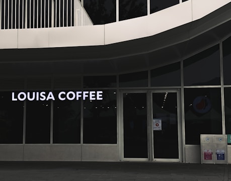 A storefront featuring large glass windows and a sign displaying 'Louisa Coffee' in bright, bold letters. The entrance consists of glass doors with signage posted on them. The exterior design is modern, with clean lines and a minimalist aesthetic. Two recycling bins are positioned to the right of the entrance.