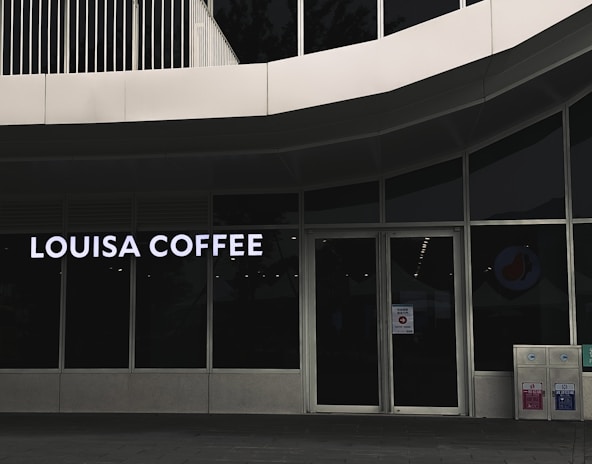 A storefront featuring large glass windows and a sign displaying 'Louisa Coffee' in bright, bold letters. The entrance consists of glass doors with signage posted on them. The exterior design is modern, with clean lines and a minimalist aesthetic. Two recycling bins are positioned to the right of the entrance.