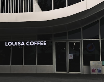 A storefront featuring large glass windows and a sign displaying 'Louisa Coffee' in bright, bold letters. The entrance consists of glass doors with signage posted on them. The exterior design is modern, with clean lines and a minimalist aesthetic. Two recycling bins are positioned to the right of the entrance.
