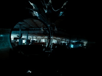 An industrial setting with dim lighting, showcasing large metal pipes and a spacious factory interior. Visible machinery and structural beams are part of the shadowy, expansive environment.