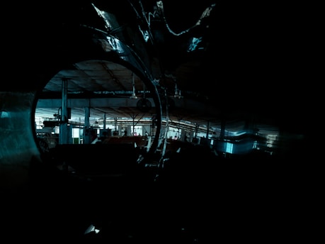 An industrial setting with dim lighting, showcasing large metal pipes and a spacious factory interior. Visible machinery and structural beams are part of the shadowy, expansive environment.