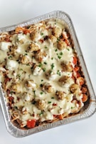 A foil tray filled with a layered pasta dish topped with creamy white sauce and meatballs, garnished with chopped green herbs. Visible layers include spiraled pasta, red tomato or pepper slices, and a generous amount of sauce.