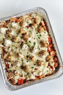 A foil tray filled with a layered pasta dish topped with creamy white sauce and meatballs, garnished with chopped green herbs. Visible layers include spiraled pasta, red tomato or pepper slices, and a generous amount of sauce.