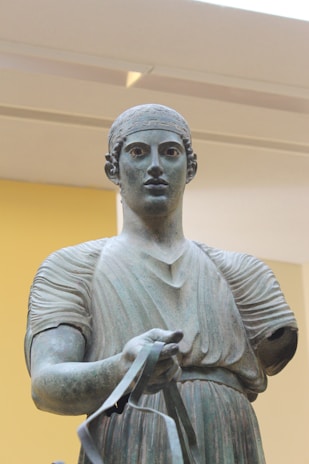 A classical bronze statue of a standing figure, possibly an ancient athlete or charioteer, displayed in an indoor setting with a soft yellow background. The figure is wearing a headband and a textured tunic, holding a metallic object in one hand, with an expressionless face.