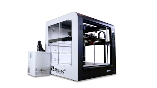 A large, modern 3D printer with a clear enclosure and a sleek design is accompanied by a separate unit. It features visible components and intricate mechanical parts within its transparent casing, highlighting its functionality.