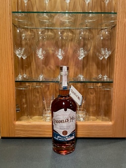 A bottle of Ruddell's Mill Kentucky Straight Bourbon Whiskey is placed in front of a wooden cabinet with glass shelves. The cabinet is filled with wine glasses and short tumblers neatly arranged on the shelves.