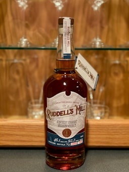 A bottle of Ruddell's Mill Kentucky Straight Bourbon Whiskey is placed on a dark surface with a wooden background. The label indicates it is a crafted small batch, with 46.1% alcohol by volume. Behind the bottle, there are glass shelves with wine glasses.