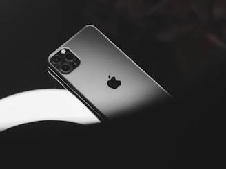A sleek, modern smartphone with a triple-camera system is resting against a glossy, dark surface. The apple logo is prominently visible on the back of the device, reflecting some light.