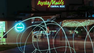 A nighttime scene at a shopping mall entrance with vibrant neon lighting. A sign reads 'Ayala Malls Central Bloc' along with a bright blue circular light on the left wall. Decorative string lights hang under a canopy, illuminating the area. Light trails swirl across the front, creating dynamic patterns.