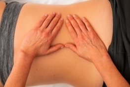 Hands are applying gentle pressure on a person's back, indicating a massage or physical therapy session. The person is lying down and wearing a gray garment.