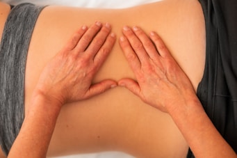 Hands are applying gentle pressure on a person's back, indicating a massage or physical therapy session. The person is lying down and wearing a gray garment.