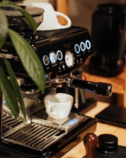 An espresso machine is placed on a wooden surface, accompanied by a white coffee cup positioned under the portafilter. The machine is sleek and modern, featuring illuminated buttons and a pressure gauge. A leafy plant is partially visible on the left side.