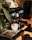 An espresso machine is placed on a wooden surface, accompanied by a white coffee cup positioned under the portafilter. The machine is sleek and modern, featuring illuminated buttons and a pressure gauge. A leafy plant is partially visible on the left side.
