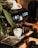 An espresso machine is placed on a wooden surface, accompanied by a white coffee cup positioned under the portafilter. The machine is sleek and modern, featuring illuminated buttons and a pressure gauge. A leafy plant is partially visible on the left side.