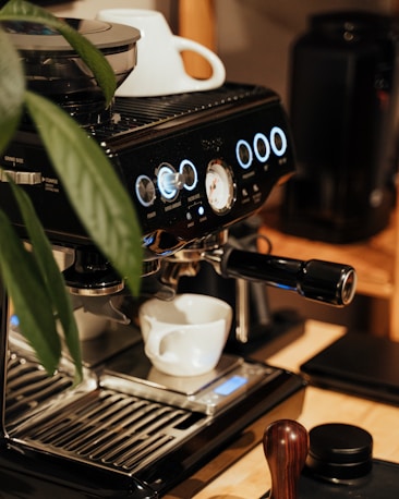 An espresso machine is placed on a wooden surface, accompanied by a white coffee cup positioned under the portafilter. The machine is sleek and modern, featuring illuminated buttons and a pressure gauge. A leafy plant is partially visible on the left side.