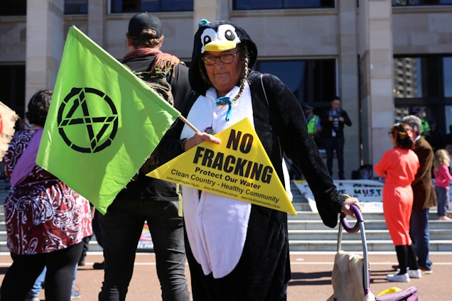A person dressed in a penguin costume holds a green flag with a black symbol and a yellow triangular sign that reads 'NO FRACKING. Protect Our Water. Clean Country - Healthy Community.' The person appears to wear glasses and a headpiece with braids. There are other people in the background, some dressed casually and others in costumes. The scene seems to take place in front of a building with steps.